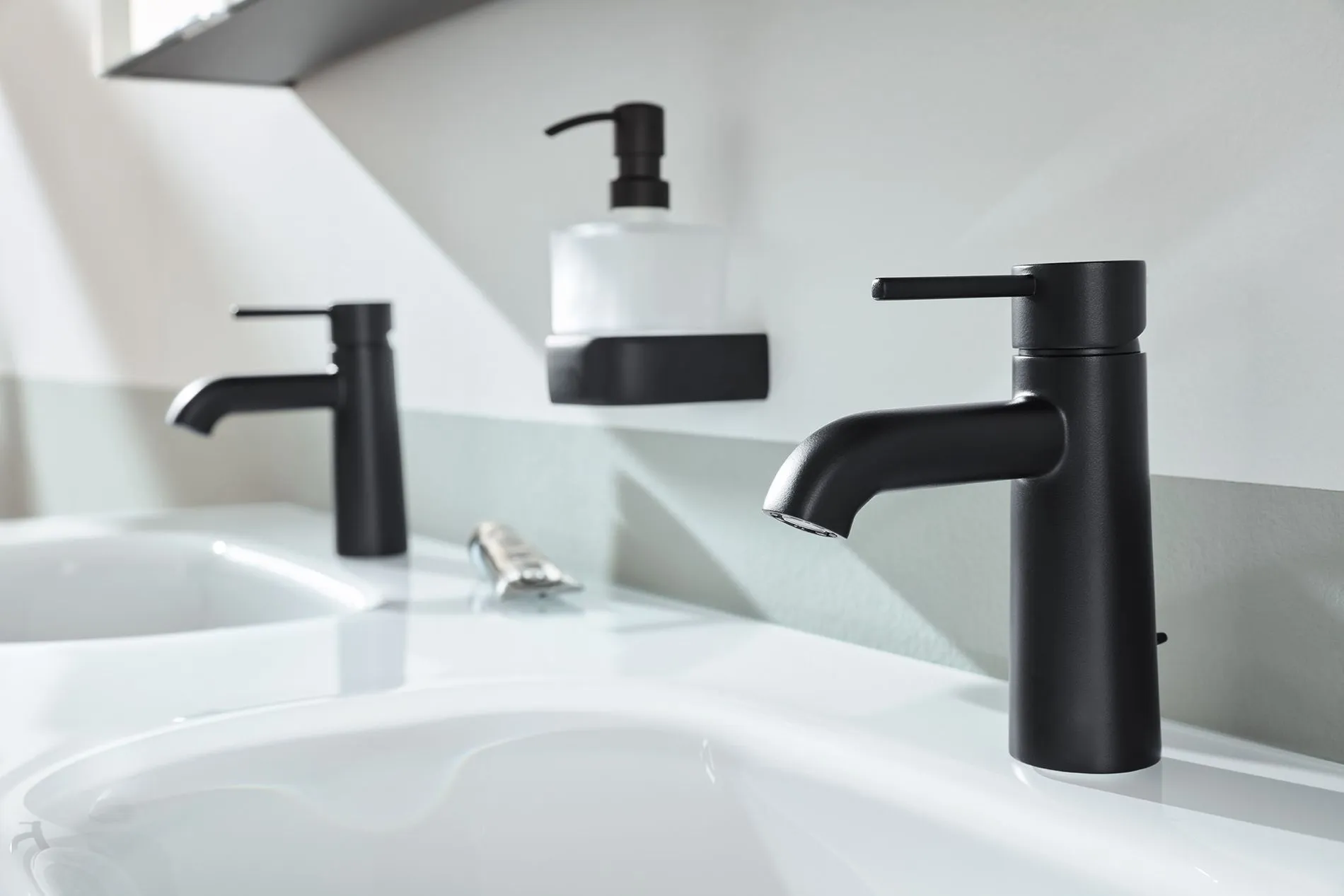 Ein modernes Waschbecken mit zwei eleganten schwarzen Wasserhähnen und einem montierten Seifenspender auf einer minimalistischen Arbeitsplatte.