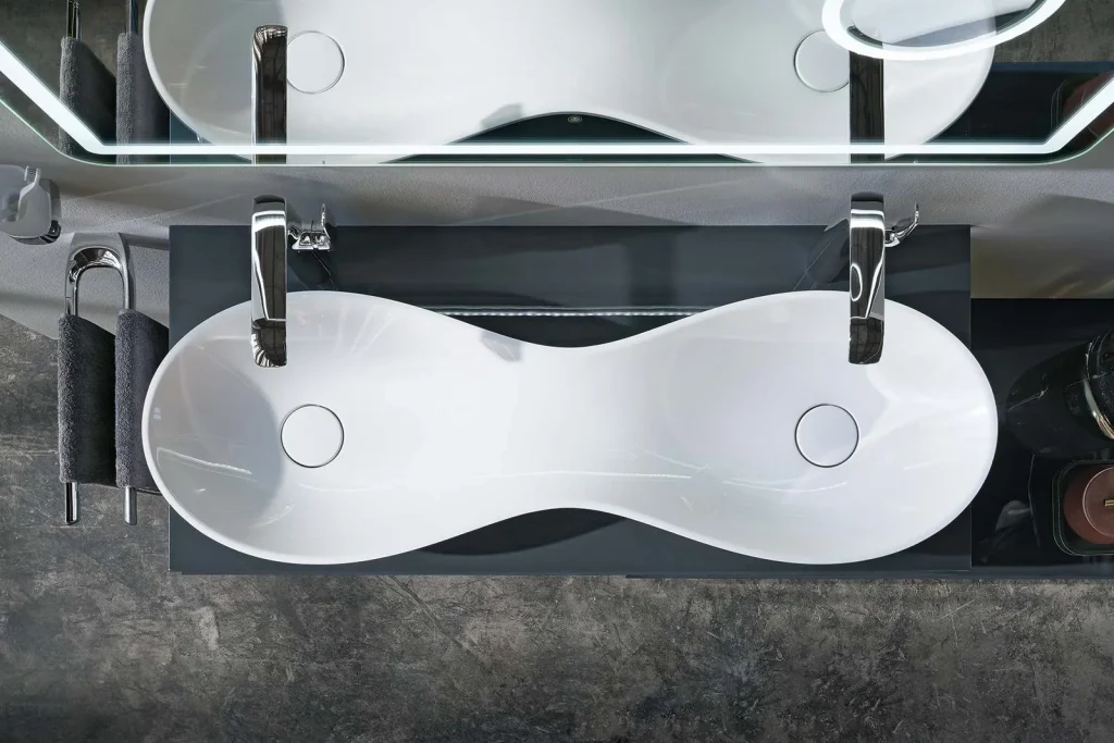 Modernes Doppelwaschbecken mit schlanker, unendlicher Form, umgeben von Chromarmaturen, schwarzem Stauraum und gerollten Handtüchern vor einem strukturierten Hintergrund.