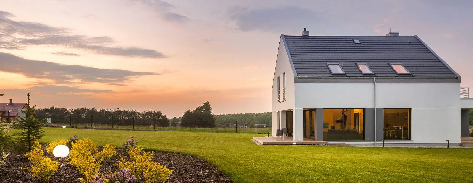Modernes weißes Haus mit dunklem Dach steht bei Sonnenuntergang auf einer weitläufigen grünen Wiese. Blumen im Vordergrund, warmes Licht aus den Fenstern und ein ruhiger Himmel.