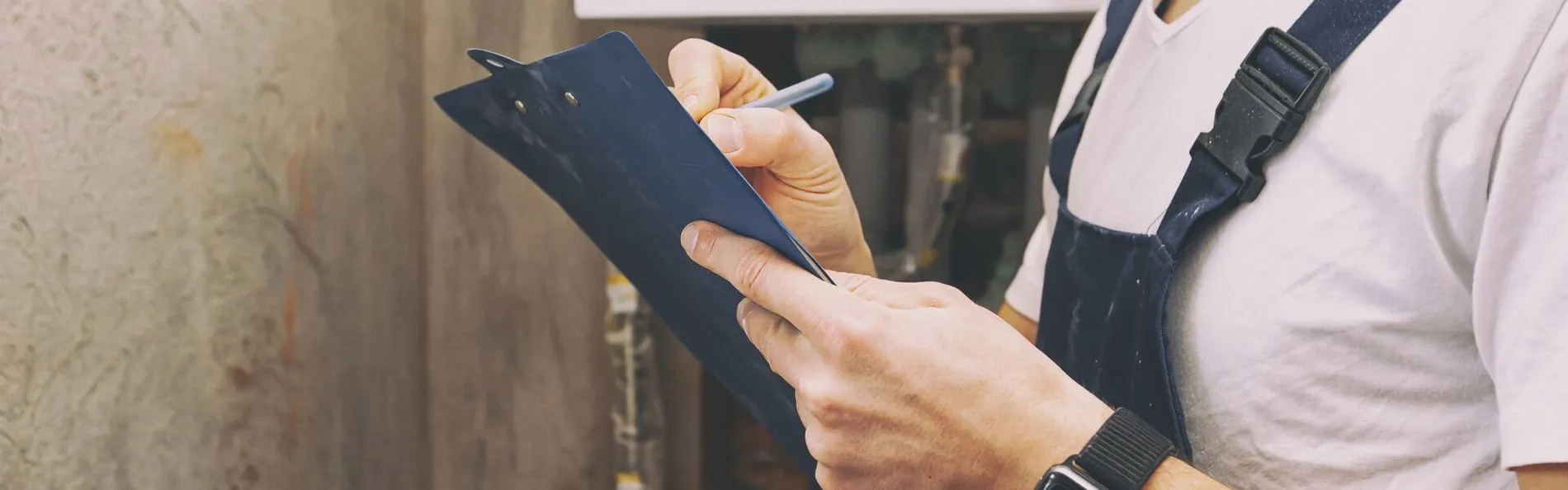 Eine Person in einem weißen T-Shirt und einem dunklen Overall hält ein blaues Klemmbrett und einen Stift in der Hand und macht sich in einer industriellen Umgebung Notizen, was Konzentration und Fleiß ausstrahlt.