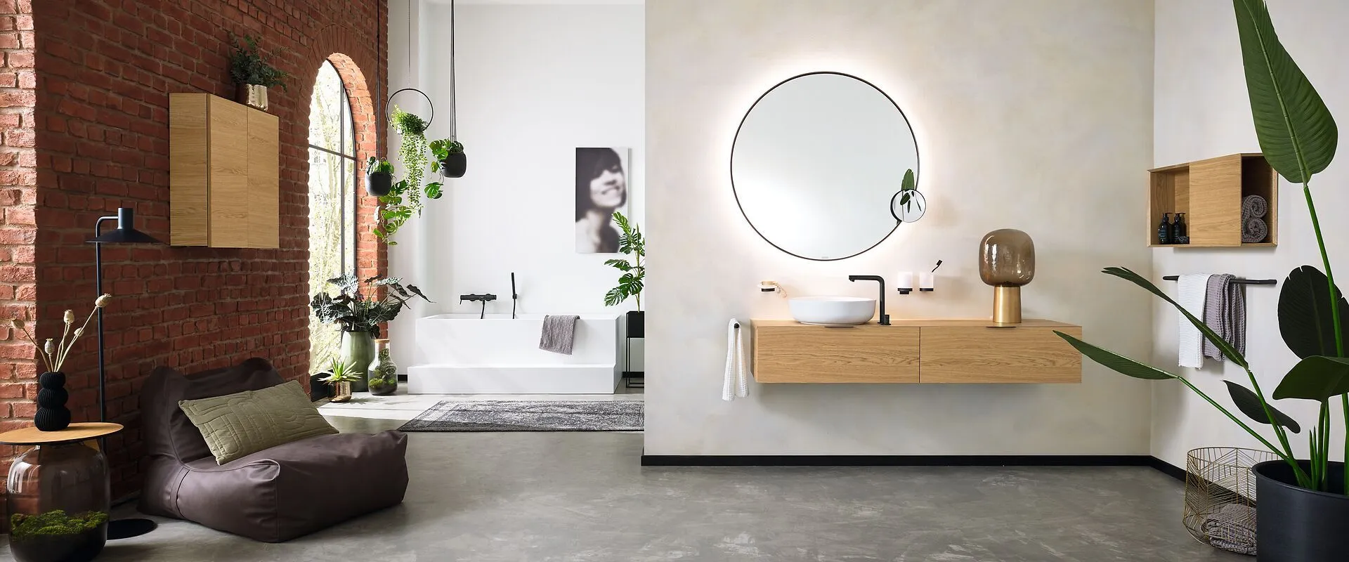 Stilvolles Badezimmer mit modernem, erdigem Design. Mit hinterleuchtetem runden Spiegel, schwebendem Holzwaschtisch, freistehender Badewanne, Ziegelwand, großen Pflanzen und sanfter Beleuchtung.